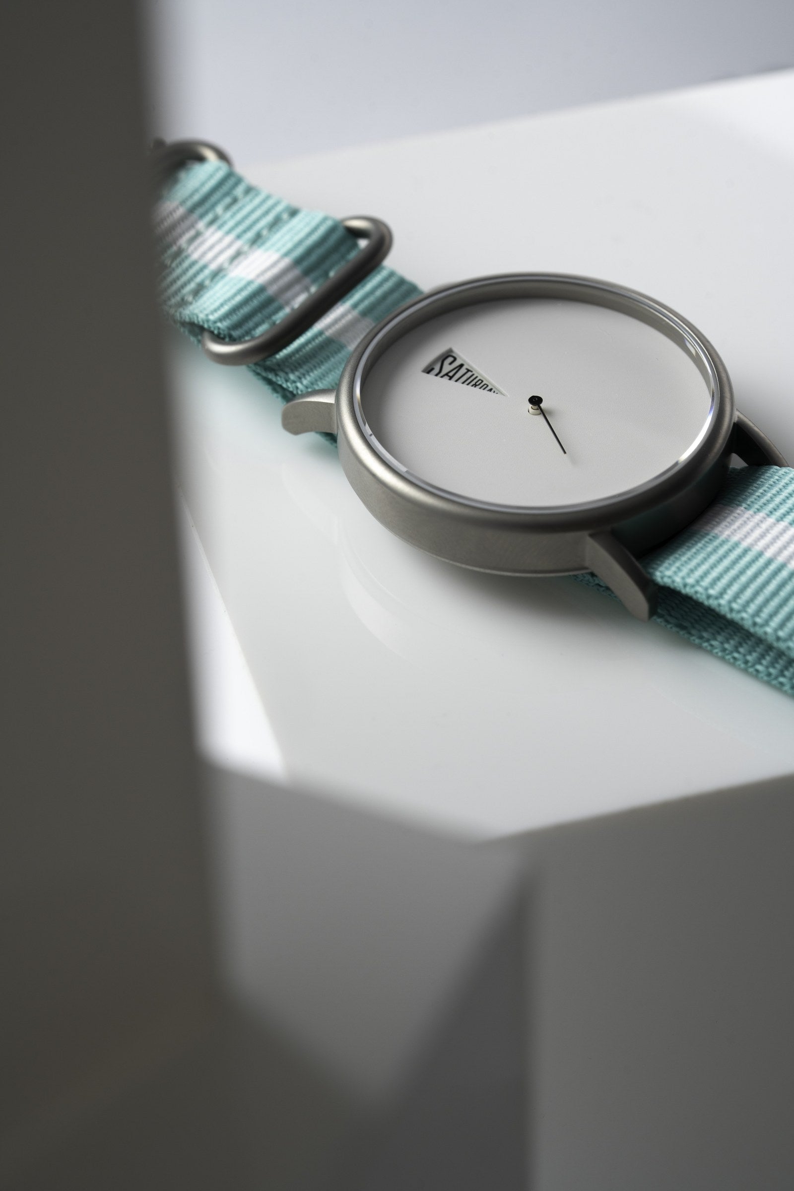 The Point White Daywatch — front face shot on geometric white surface with teal NATO strap