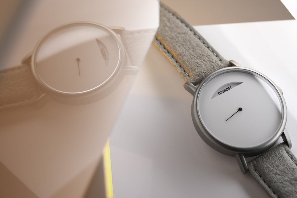 The Wednesday by Daywatch — two watches on warm surface, grey suede strap version in focus showing THURSDAY