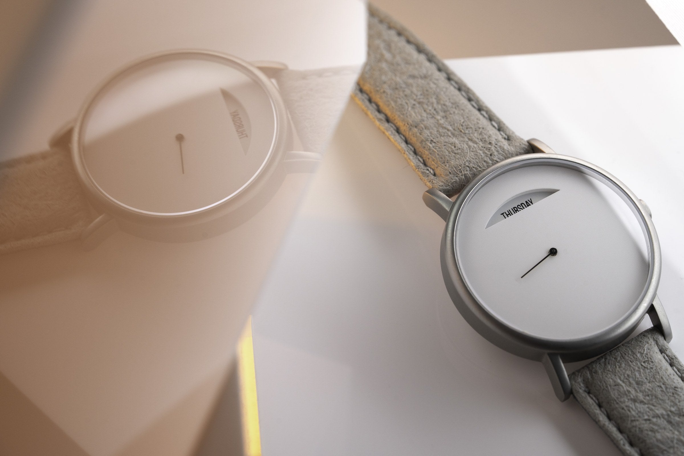The Wednesday by Daywatch — two watches on warm surface, grey suede strap version in focus showing THURSDAY